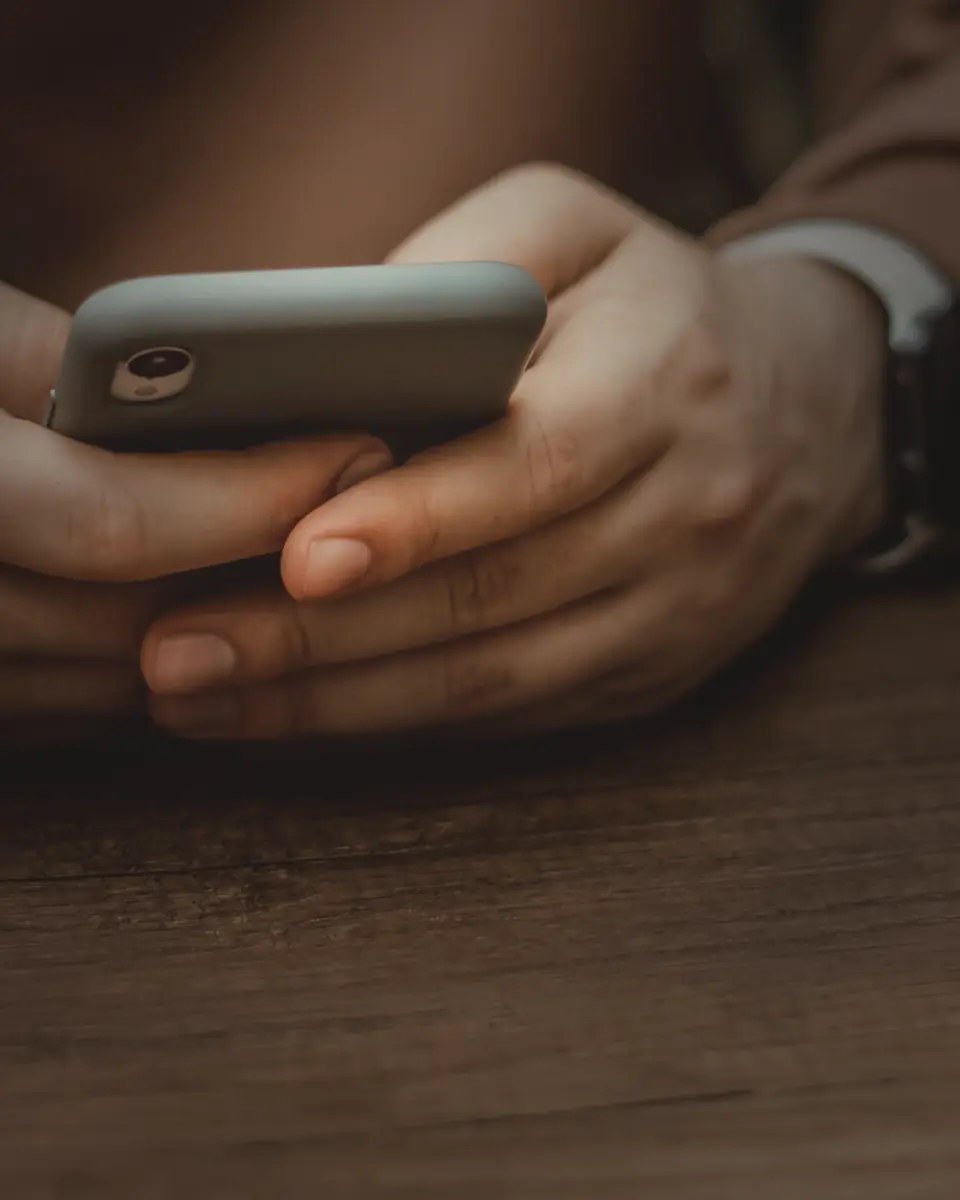 A man’s hands holding a phone at a wooden table in soft light.