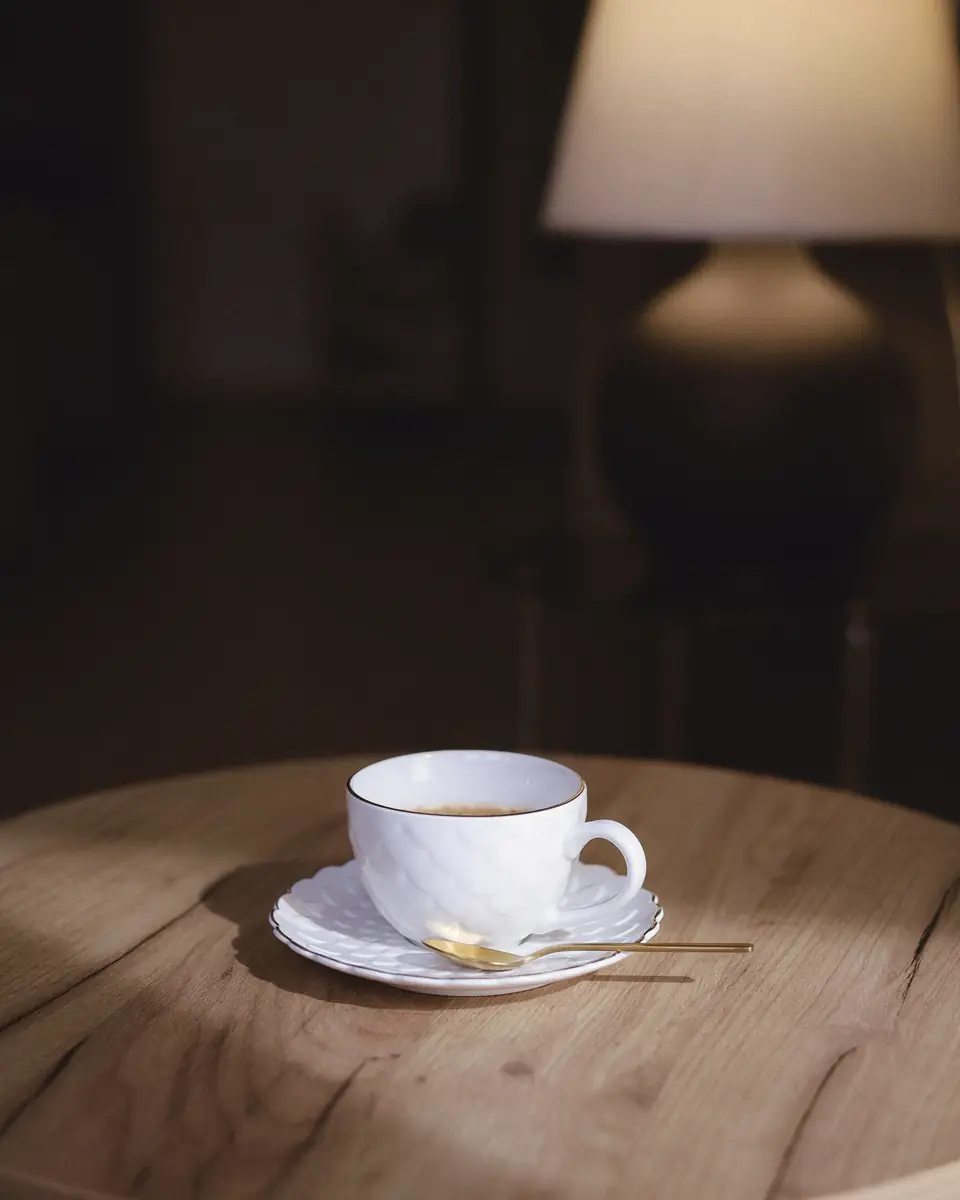 A table lamp switched on behind a cup of coffee on a small wooden table.